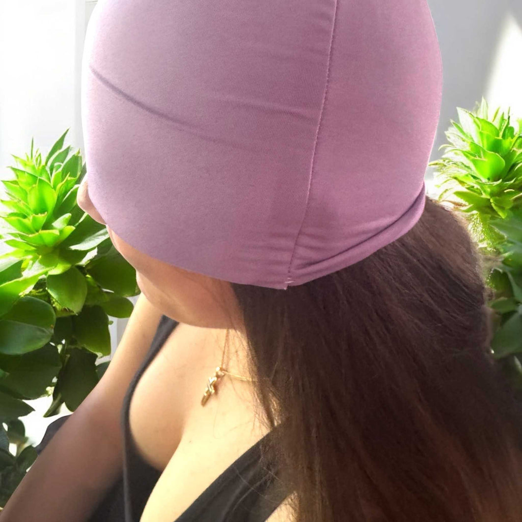 Woman wearing a mauve soothing cooling cap for migraine and headache relief surrounded by green plants