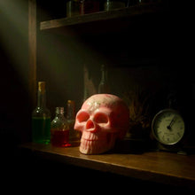 Charger l&#39;image dans la galerie, Red glycerin soap shaped like a skull on a wooden shelf with vintage bottles and a clock in dim lighting
